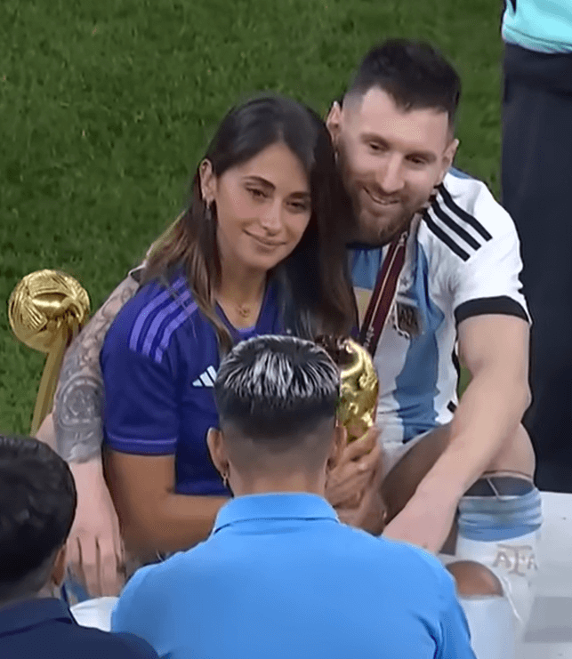 Lionel Messi abrazando a su esposa Antonela Roccuzzo mientras sostiene el trofeo de la Copa del Mundo 2022, celebrando en el campo tras la victoria de Argentina.